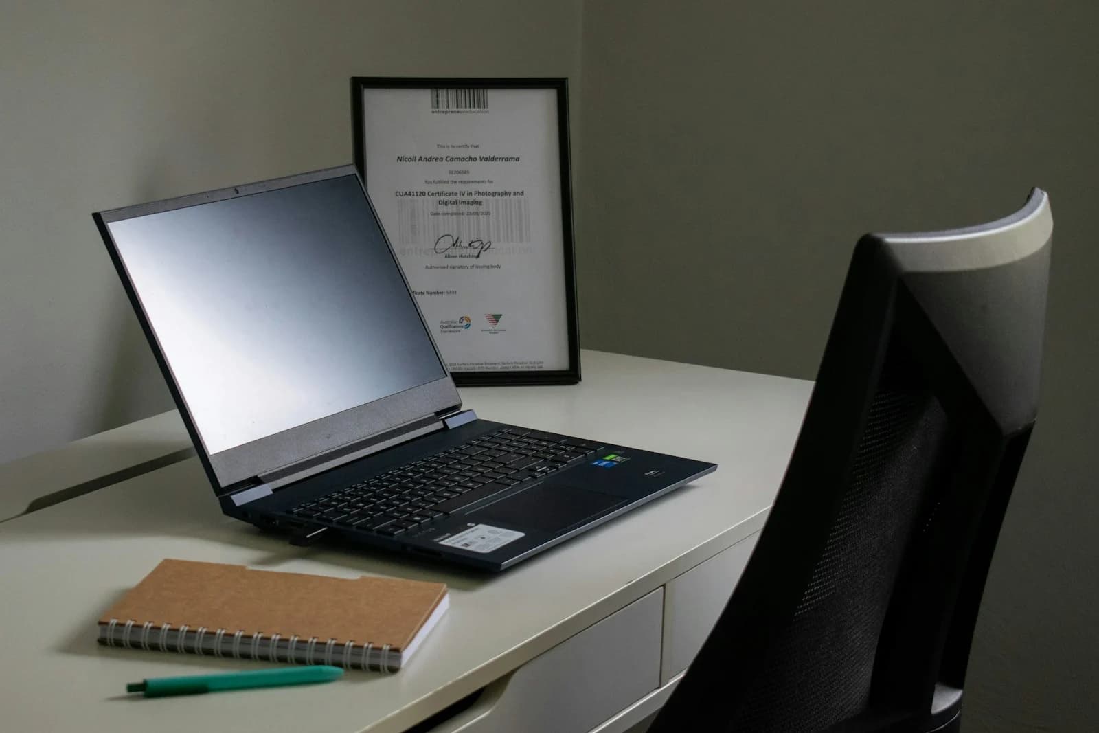 An open laptop and a small notebook on a tidy desk beside a chair.
