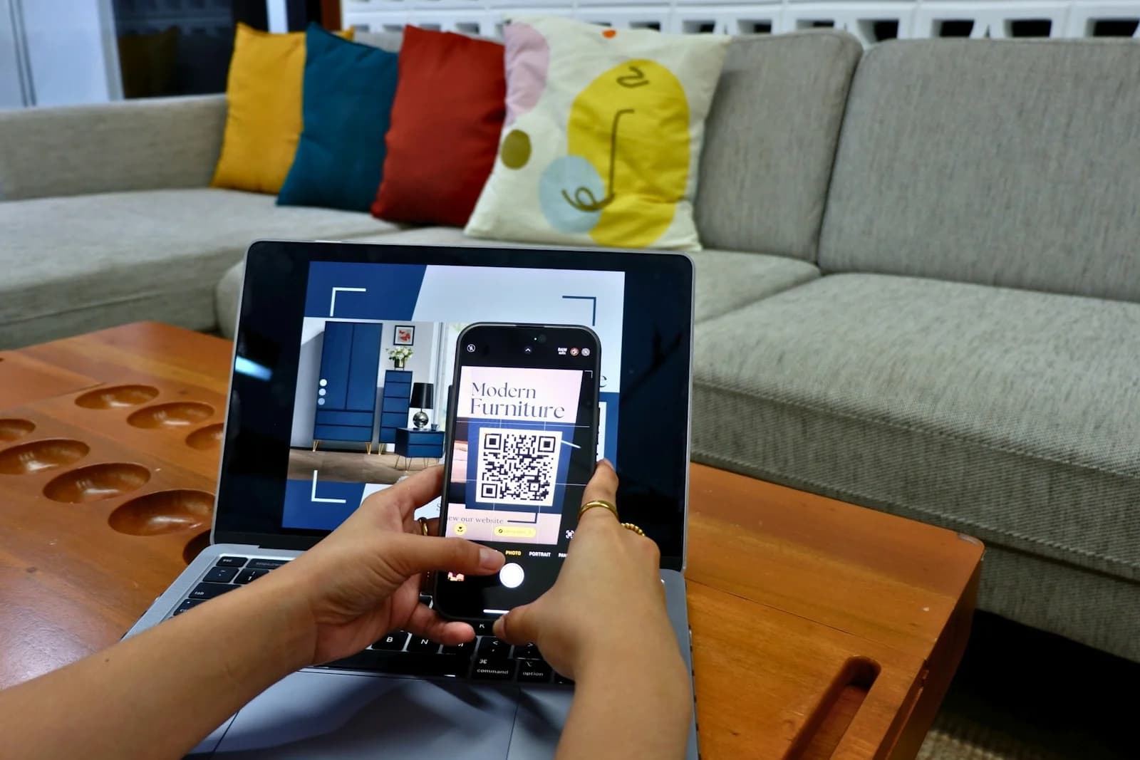 A person holds a phone in front of a QR code displayed on a laptop at a coffee table.
