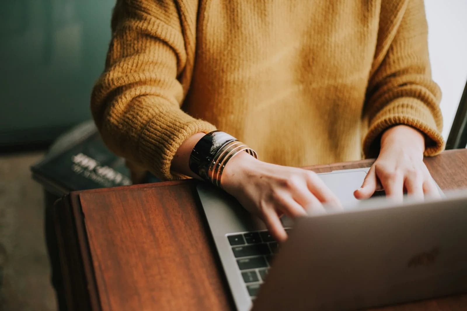 A person in a mustard sweater works on a laptop at a wooden table.