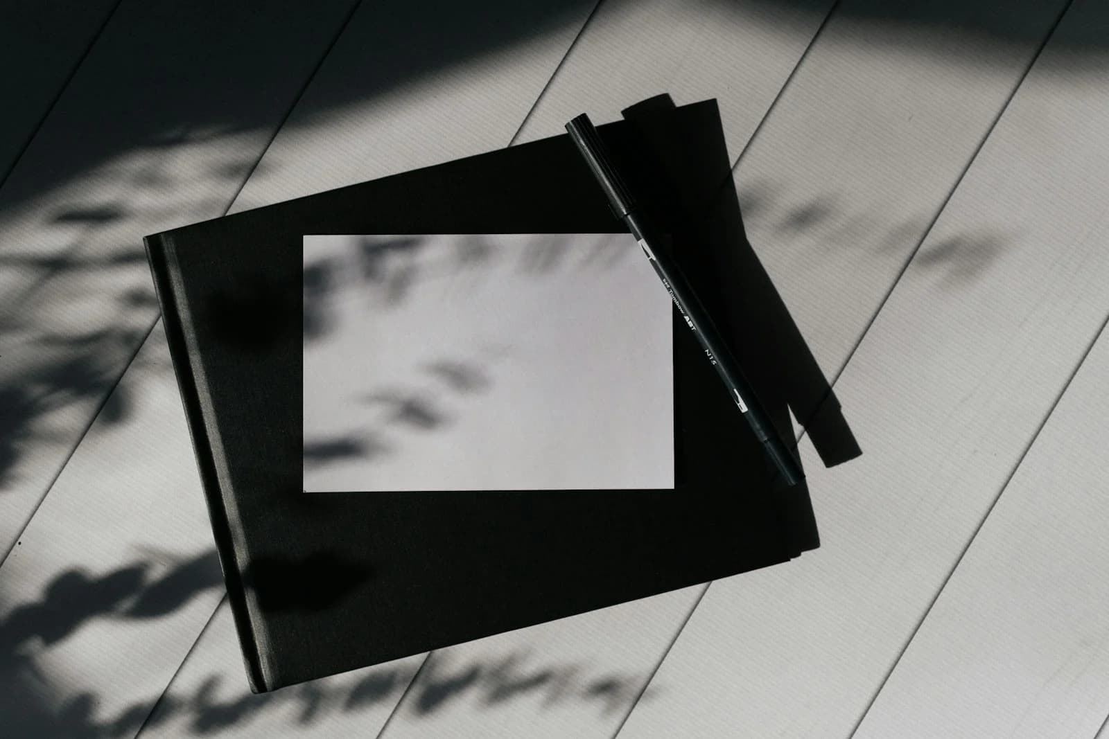 A black pen beside a blank notebook on a pale desk, with soft leaf shadows.