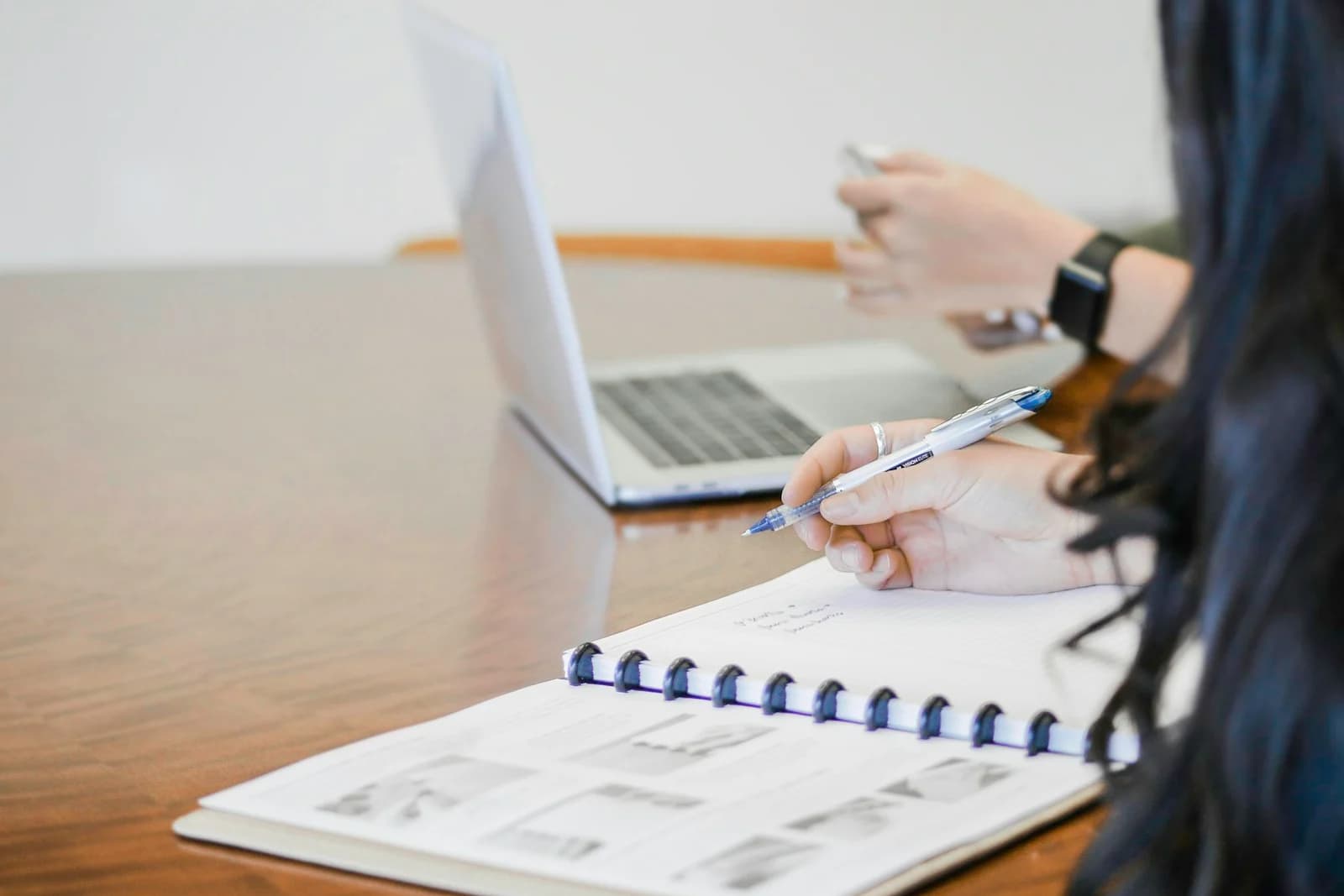A person writing in a notebook at a meeting table beside a laptop.